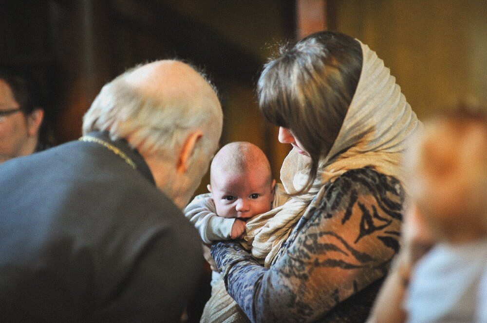 Mutter hält ihr Baby im Arm, während der Priester die Taufzeremonie fortsetzt