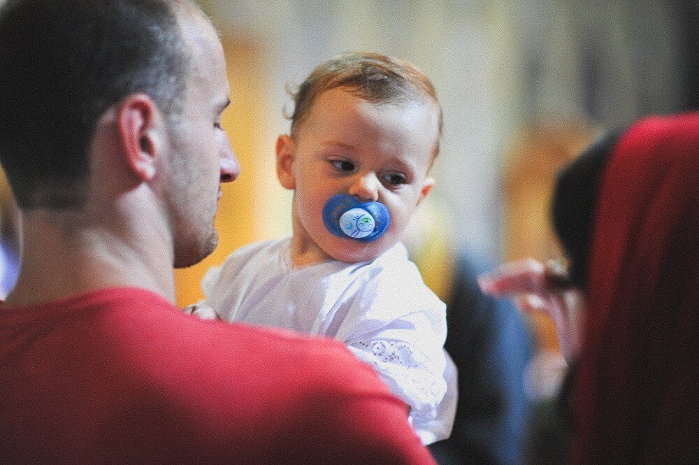 Vater hält ein Baby im weißen Taufgewand mit blauem Schnuller