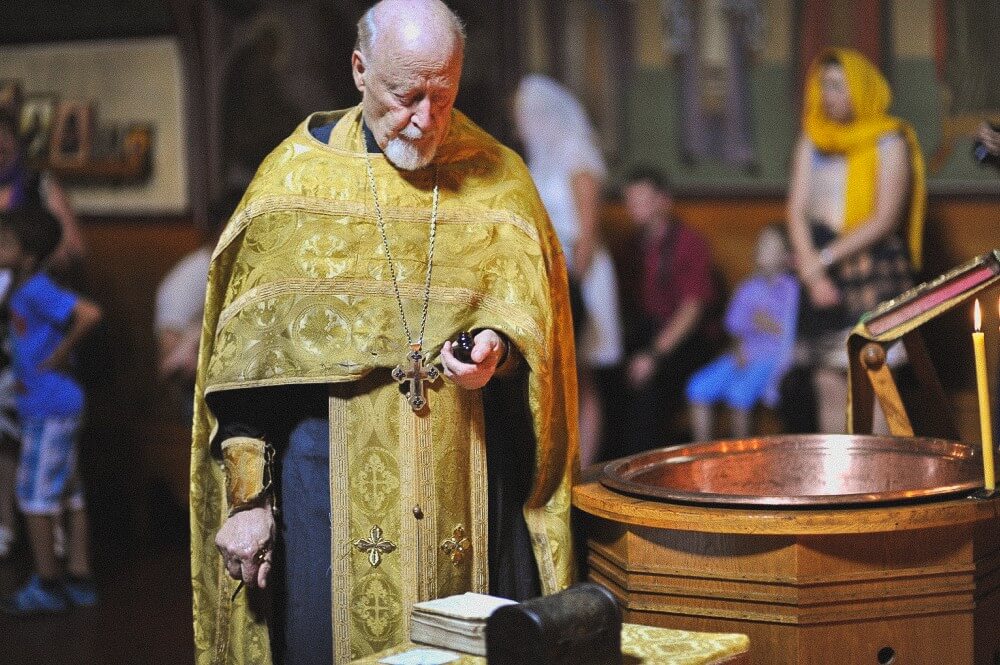 Priester steht mit Weihöl und liturgischen Gegenständen neben dem Taufbecken